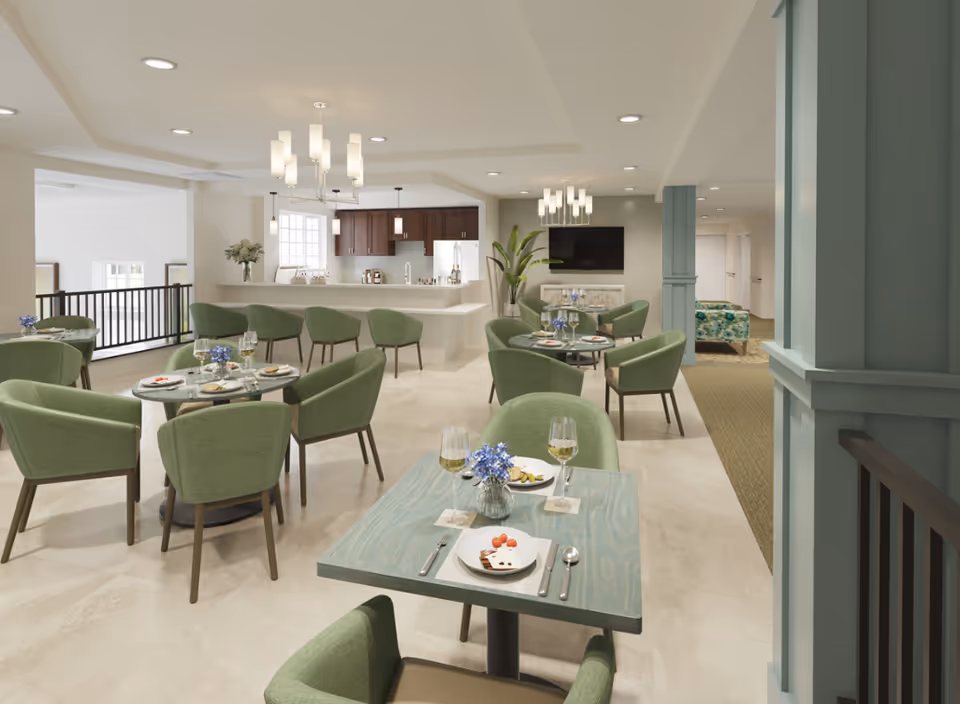 A bright and modern dining area in a senior living facility featuring multiple tables with green upholstered chairs. Each table is set with plates, glasses of white wine, and small flower arrangements. In the background, there is a kitchen area with dark wood cabinets, a white refrigerator, and a countertop with bar stools. The room is well-lit with ceiling lights and chandeliers, and there is a TV mounted on the wall with a plant beside it.