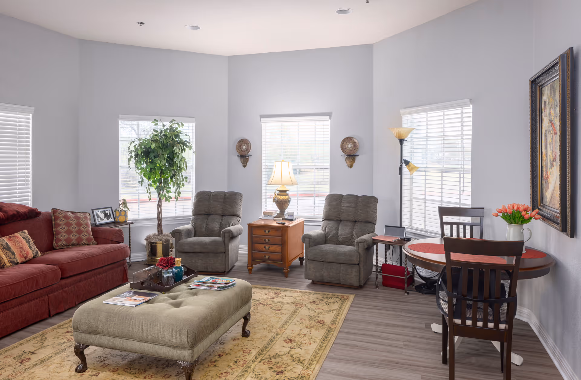 A bright and cozy living room with a red sofa, two gray recliner chairs, a large cushioned ottoman, a wooden side table with a lamp, a small round dining table with two chairs, a floor lamp, a potted plant, and a framed painting on the wall. The room has large windows with white blinds and light gray walls.