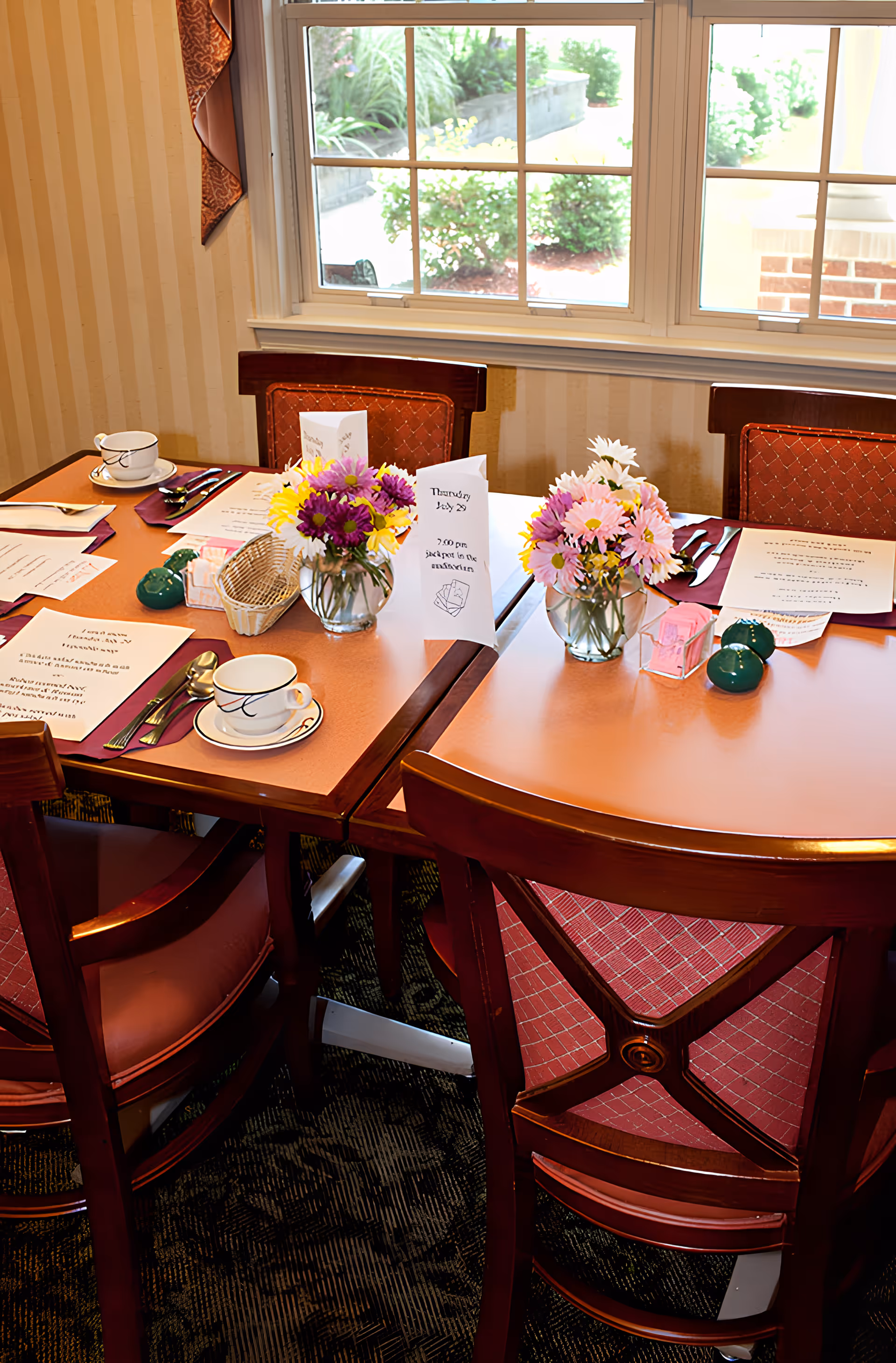 A dining table set for a meal with four wooden chairs around it. The table has two small vases with colorful flowers, menus or event cards, cups and saucers, utensils, and salt and pepper shakers. A window behind the table shows greenery outside.