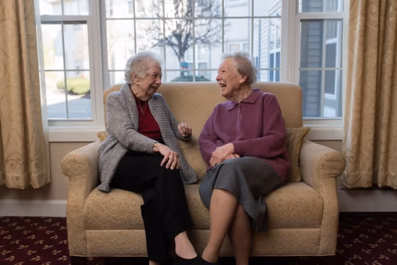 Two elderly women sitting on a beige patterned loveseat in a well-lit room with large windows behind them, smiling and engaged in conversation. The room has cream-colored curtains and a dark carpet with a subtle pattern.