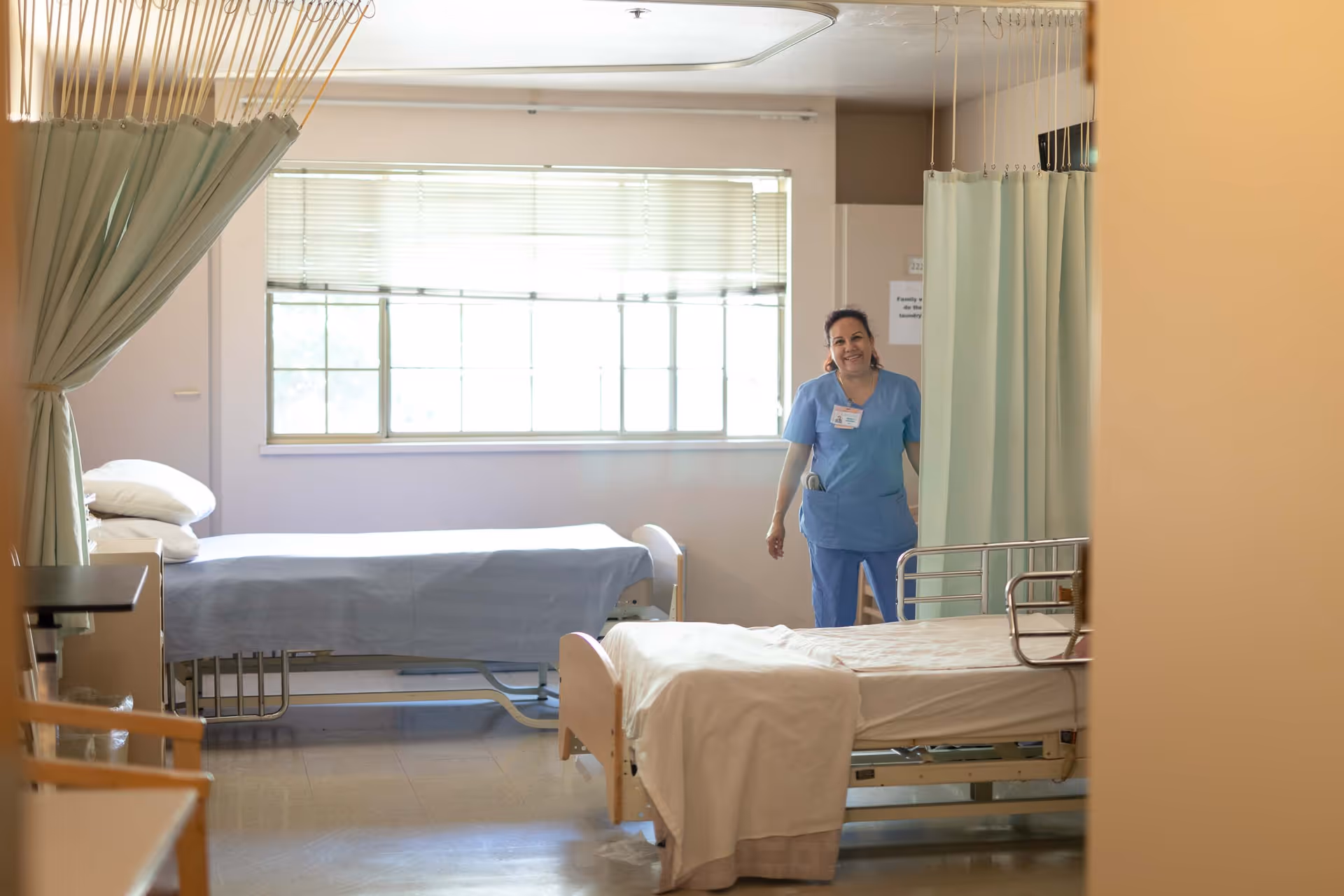 A nursing room with two hospital beds separated by curtains. A smiling healthcare worker in blue scrubs stands near the window in the background. The room is well-lit with natural light coming through a large window with blinds.