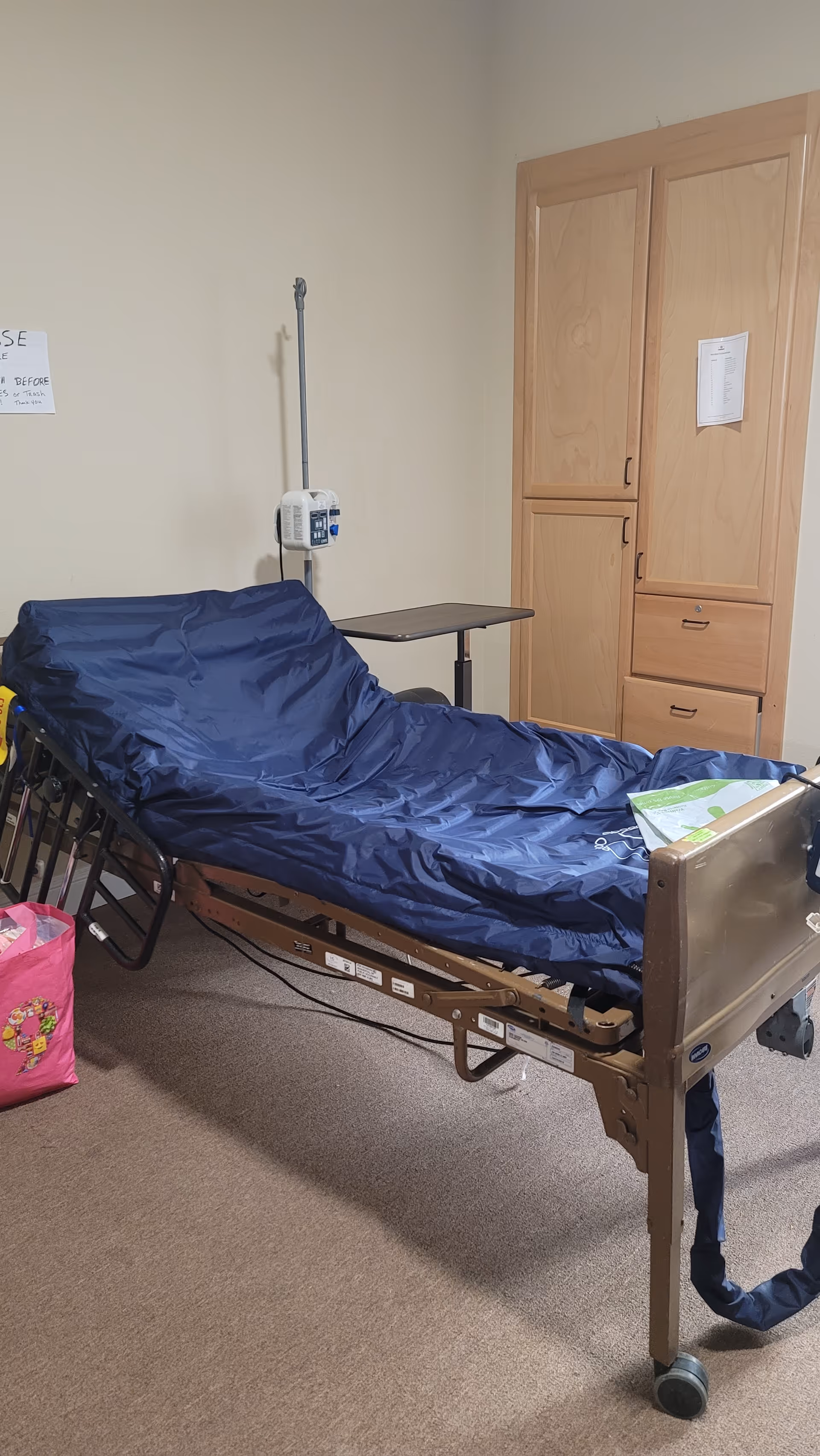 A hospital-style adjustable bed with a navy cover in a small resident bedroom, next to an overbed table and wooden wardrobe.
