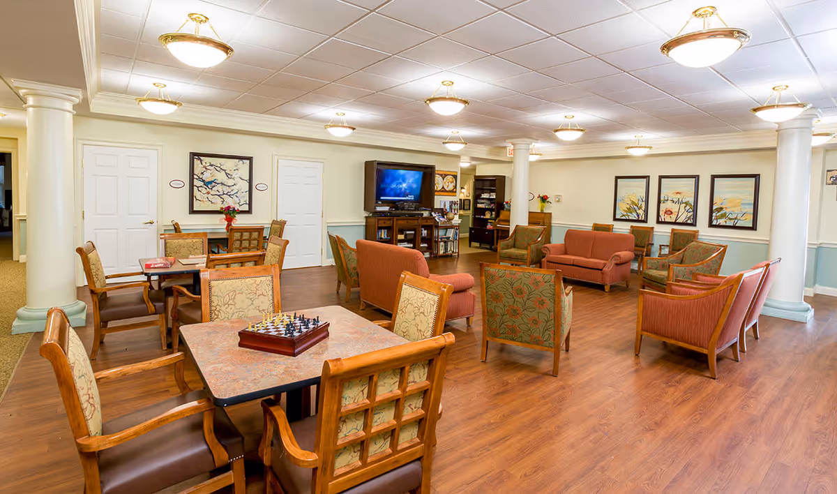 A spacious senior living common area with multiple seating arrangements including armchairs and sofas in warm tones. There are several tables with chairs, one of which has a chessboard set up. The room features wood flooring, white columns, and soft overhead lighting. Artwork is displayed on the walls, and a television is mounted on a wooden cabinet in the background.
