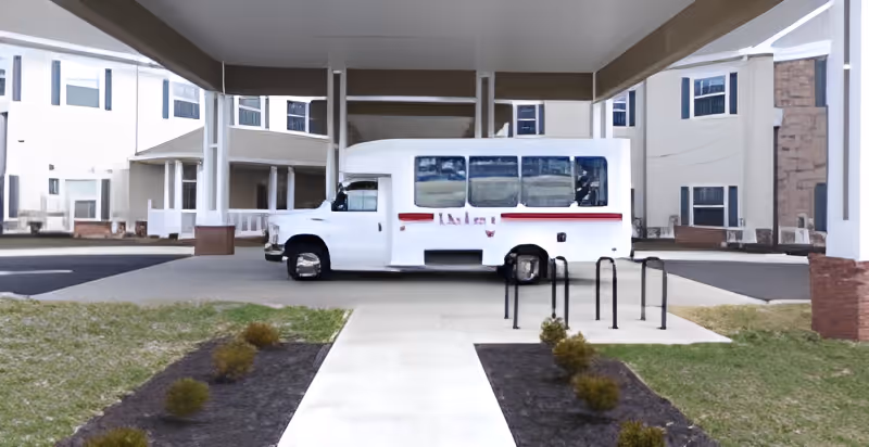 A white passenger shuttle van parked beneath a covered entrance/porte-cochère in front of a multi-story senior living building.