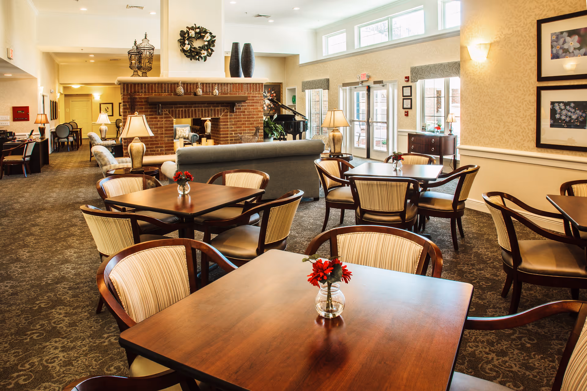 A spacious and well-lit common area in a senior living facility featuring several wooden tables with cushioned chairs arranged around them. Each table has a small vase with red flowers. In the background, there is a cozy seating area with sofas and armchairs around a brick fireplace. Large windows and glass doors allow natural light to fill the room. The walls are decorated with framed artwork and a wreath above the fireplace.