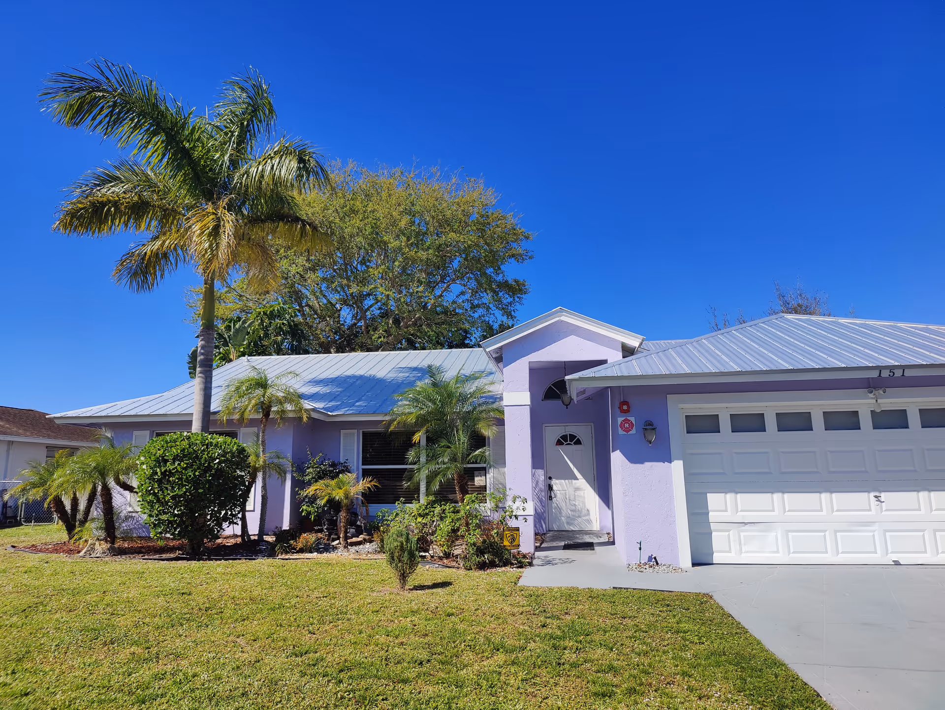 Exterior view of a single-story light purple house with a metal roof, a white garage door, and a white front door. The front yard has a well-maintained lawn, several palm trees, and other green shrubs under a clear blue sky.