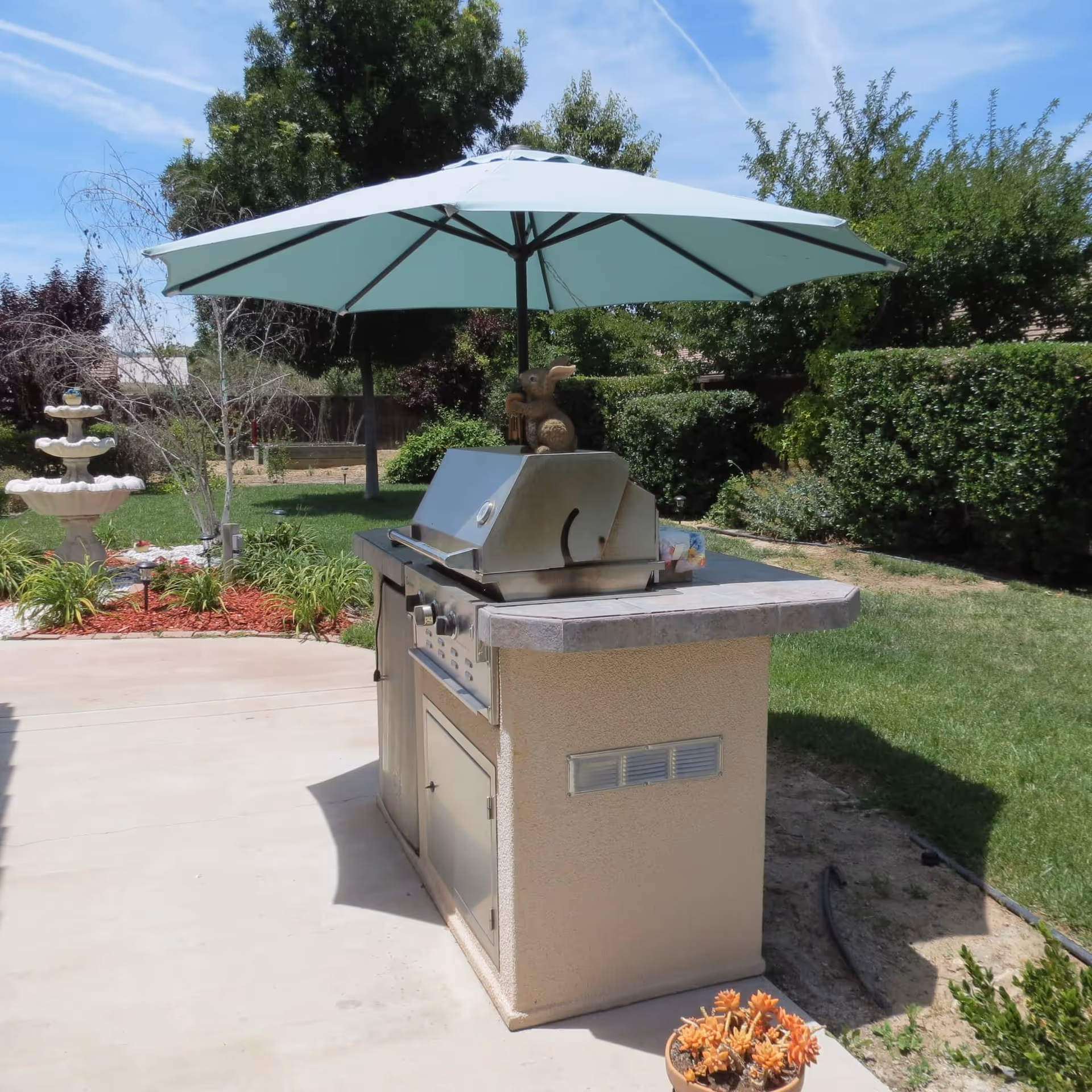 Outdoor built-in grill with a turquoise patio umbrella on a concrete patio, with lawn, hedges, and a tiered fountain in the background.