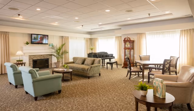 A spacious, well-lit living room with beige carpet and cream-colored walls. The room features a green sofa, two green armchairs, a beige armchair, a round wooden coffee table with a plant and decorative items, a fireplace with a TV mounted above it, a grand piano, a tall wooden grandfather clock, and a round dining table with four chairs near large windows with sheer curtains.