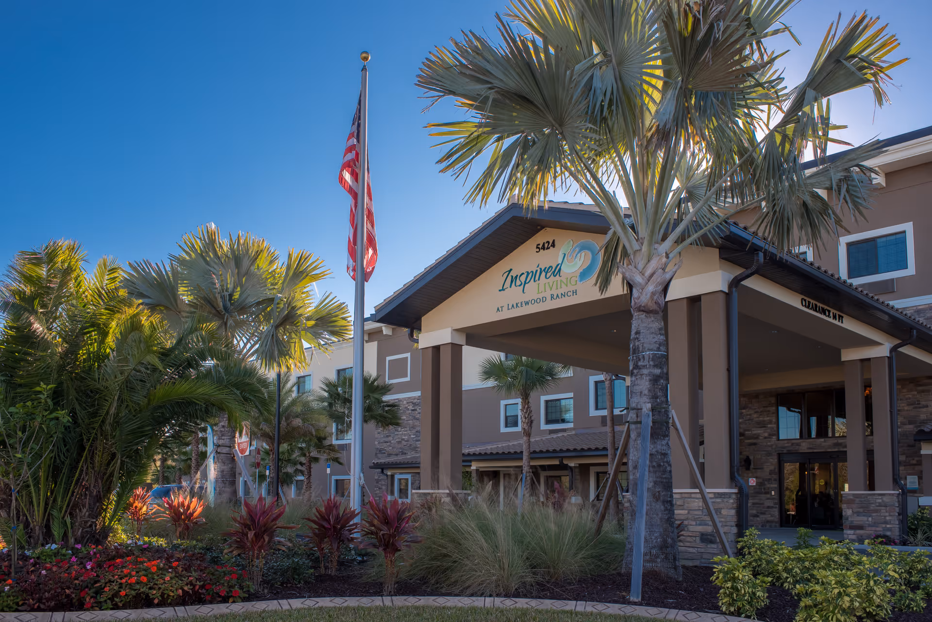 Exterior view of the entrance to Inspired Living at Lakewood Ranch, a senior living facility, featuring a covered driveway with the facility's name and address displayed. The scene includes palm trees, landscaped plants, and an American flag on a flagpole against a clear blue sky.