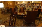 A cozy library or reading room with several wooden chairs and armchairs arranged around a central coffee table. The room has bookshelves filled with books along the back wall, framed pictures on the walls, and warm lighting creating a comfortable atmosphere.