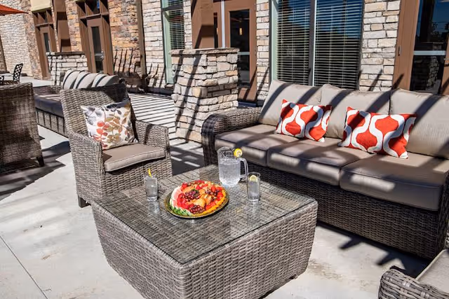 Outdoor patio area with wicker furniture including a sofa with three red and white patterned pillows, an armchair with a floral pillow, and a glass-top coffee table holding a pitcher of water, two glasses with lemon slices, and a plate of assorted fruit. The background shows a stone and brick building exterior with windows and shadows cast by the sun.
