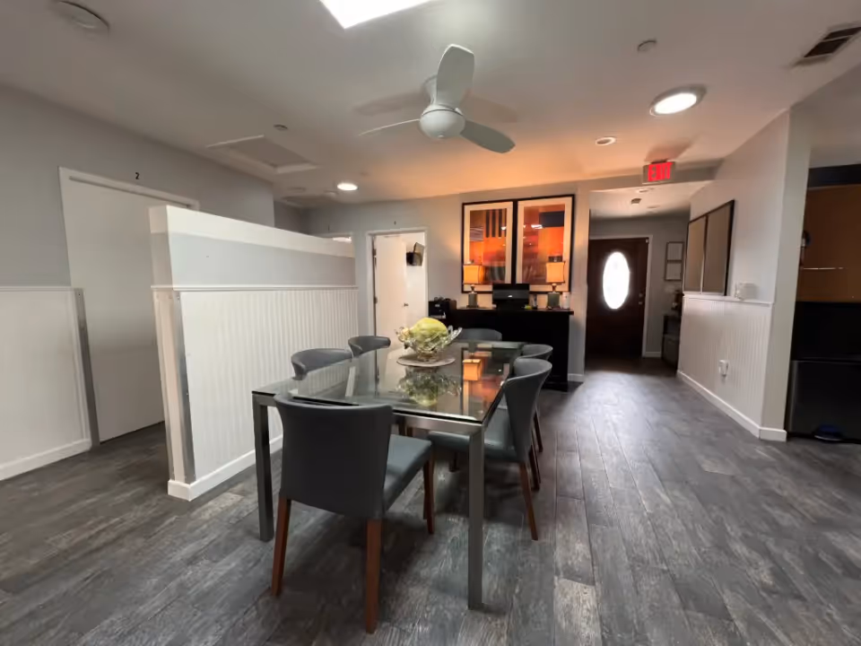 Interior view of a dining area with a glass-top table surrounded by six gray chairs. The room has gray wood flooring, white walls with wainscoting, and a ceiling fan. There are two framed artworks on the far wall above a black sideboard with two lamps. A door with an oval window is visible in the background.