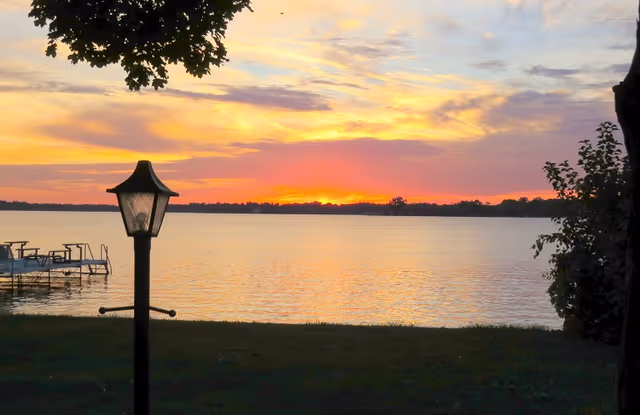 Sunset over a calm lake with a small dock to the left and a lamppost in the foreground framed by tree branches.