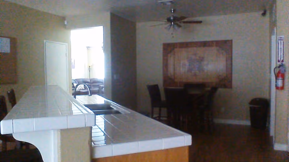 Open communal kitchen with a tiled breakfast bar and sink, bar stools, a dining table and chairs, and a seating area visible through a doorway.