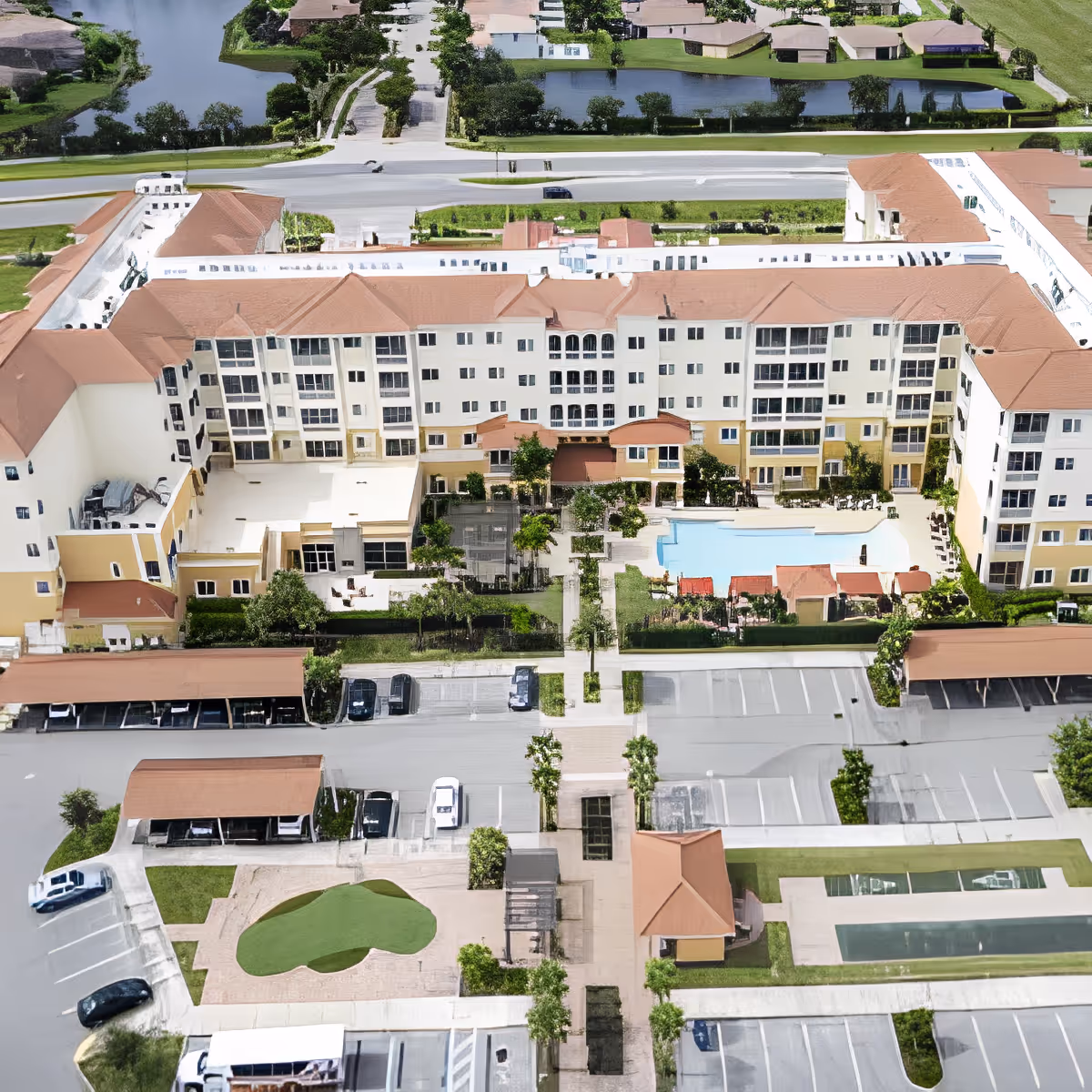 Aerial view of Discovery Village At The Forum senior living facility showing a large multi-story building with a central courtyard featuring a swimming pool, landscaped gardens, and seating areas. Surrounding the building are parking lots, covered parking spaces, and a small putting green. The facility is bordered by roads and water bodies with residential houses in the background.