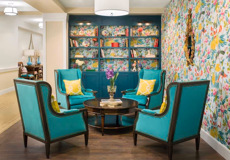 A cozy seating area with four teal armchairs arranged around a round wooden coffee table with a vase of purple flowers and books. Behind the chairs is a built-in bookshelf with floral wallpaper and various decorative items and books. The right wall also features colorful floral wallpaper and a gold-framed mirror.