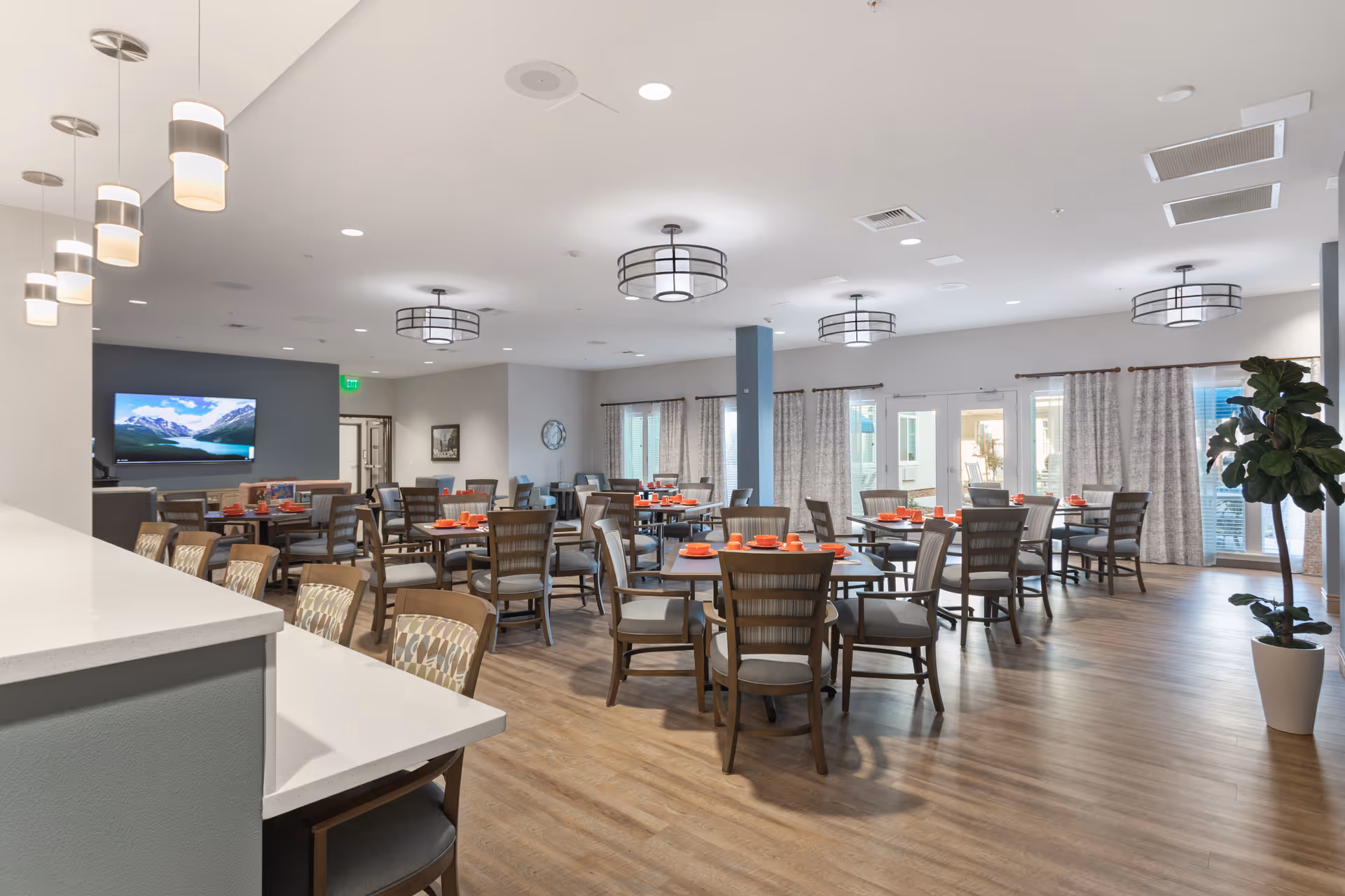 A spacious dining room with multiple round and rectangular tables surrounded by wooden chairs. Each table is set with red plates and cups. The room has large windows with curtains, modern ceiling lights, and a flat-screen TV mounted on a dark accent wall. The floor is wood, and there is a white countertop with bar stools in the foreground.