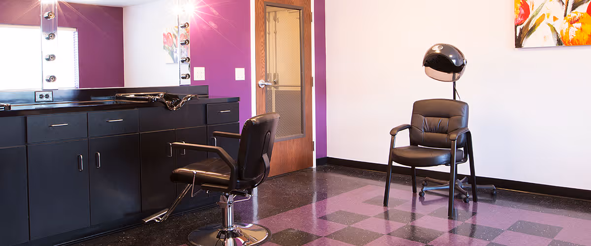 Interior of a salon room with two black salon chairs, one with a hair dryer hood attached. The room has black and purple checkered flooring, a purple accent wall with a large mirror and black cabinets, and a wooden door with a glass panel.