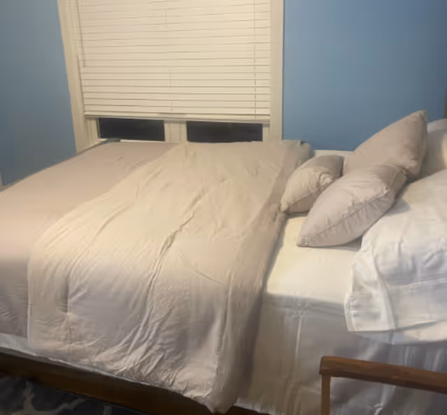 A neatly made bed with beige bedding and multiple pillows against a light blue wall. A window with closed white blinds is above the bed, and part of a wooden chair is visible at the bottom right corner.