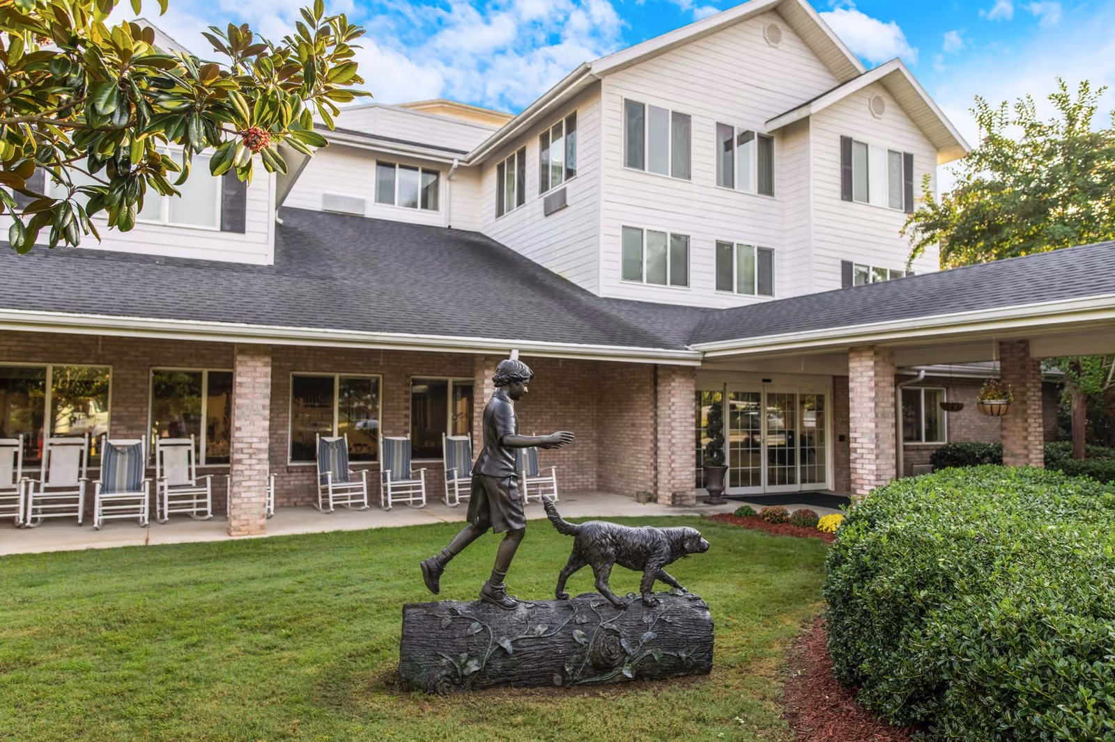 Front entrance of a multi-story senior living building with a porch of rocking chairs and a bronze statue of a child walking a dog on the lawn.