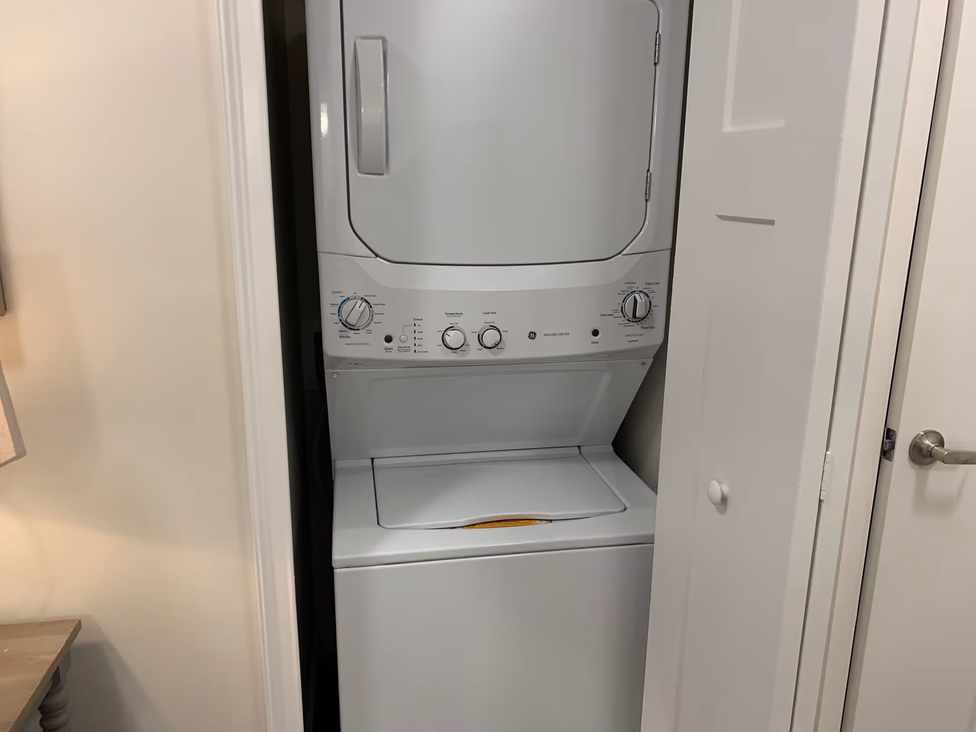 A stacked white washer and dryer unit installed in a small laundry closet with white walls and a partially open white door.
