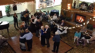 A group of elderly people dancing and socializing in a warmly lit common area with a fireplace, string lights, and a live band playing music. Some people are seated at tables, while others are on the dance floor.