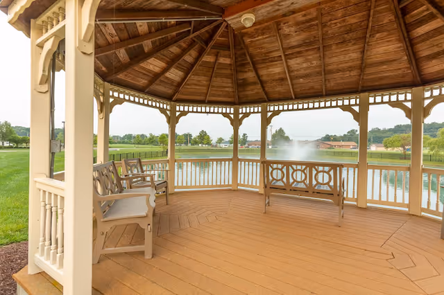 An open wooden gazebo with benches overlooking a pond and fountain on a grassy lawn.