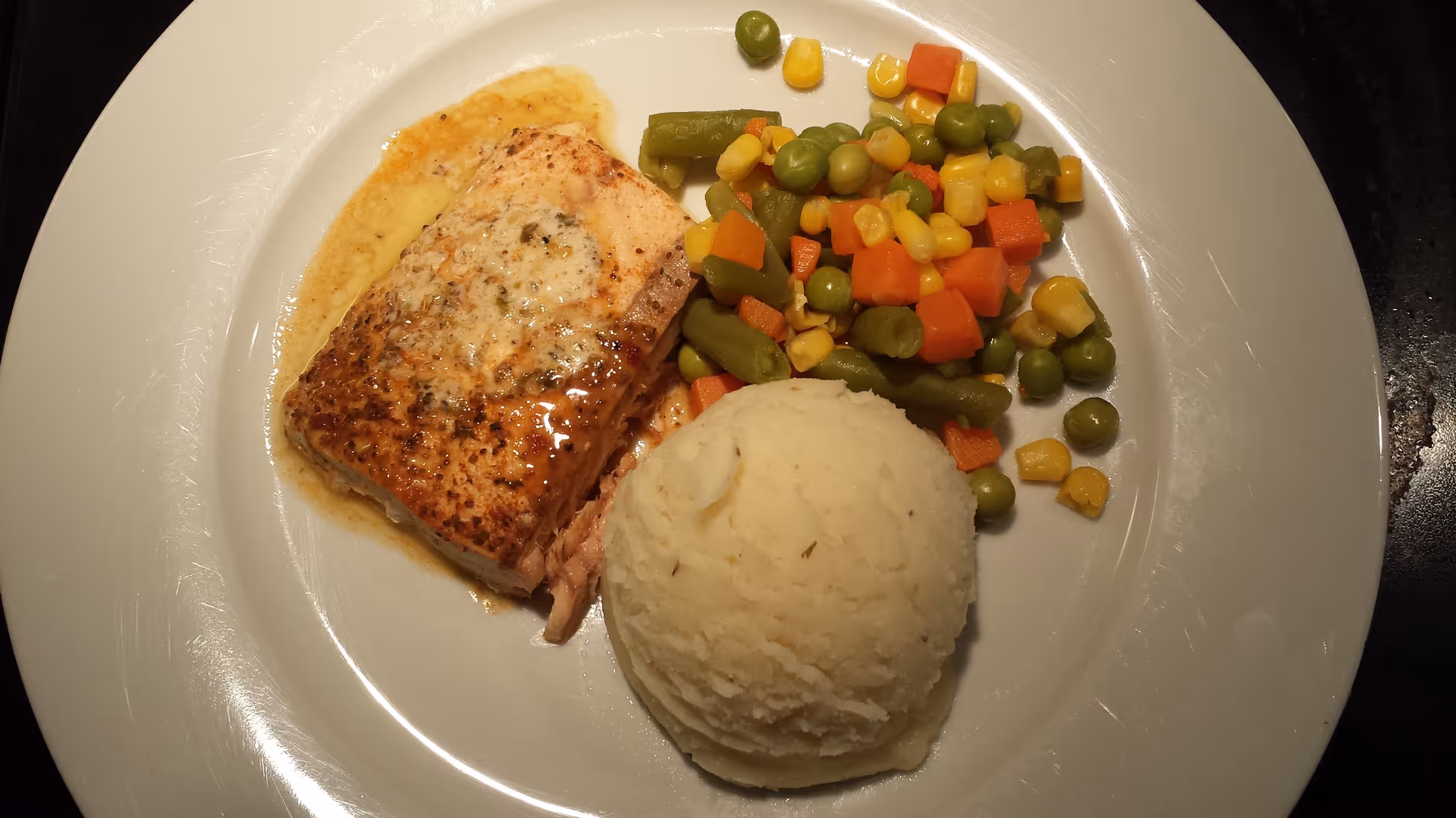 A white plate with a serving of cooked salmon topped with a light sauce, a scoop of mashed potatoes, and a mixed vegetable medley including green beans, peas, corn, and diced carrots.