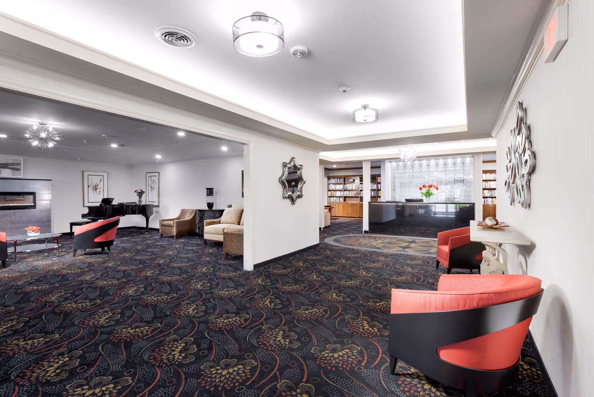 A spacious, well-lit common area in a senior living facility featuring patterned carpet, modern ceiling lights, and comfortable seating including red and beige chairs. The room includes a black grand piano, a fireplace, framed artwork on the walls, and a reception desk with bookshelves behind it. Decorative mirrors and a small table with a flower vase are also visible.