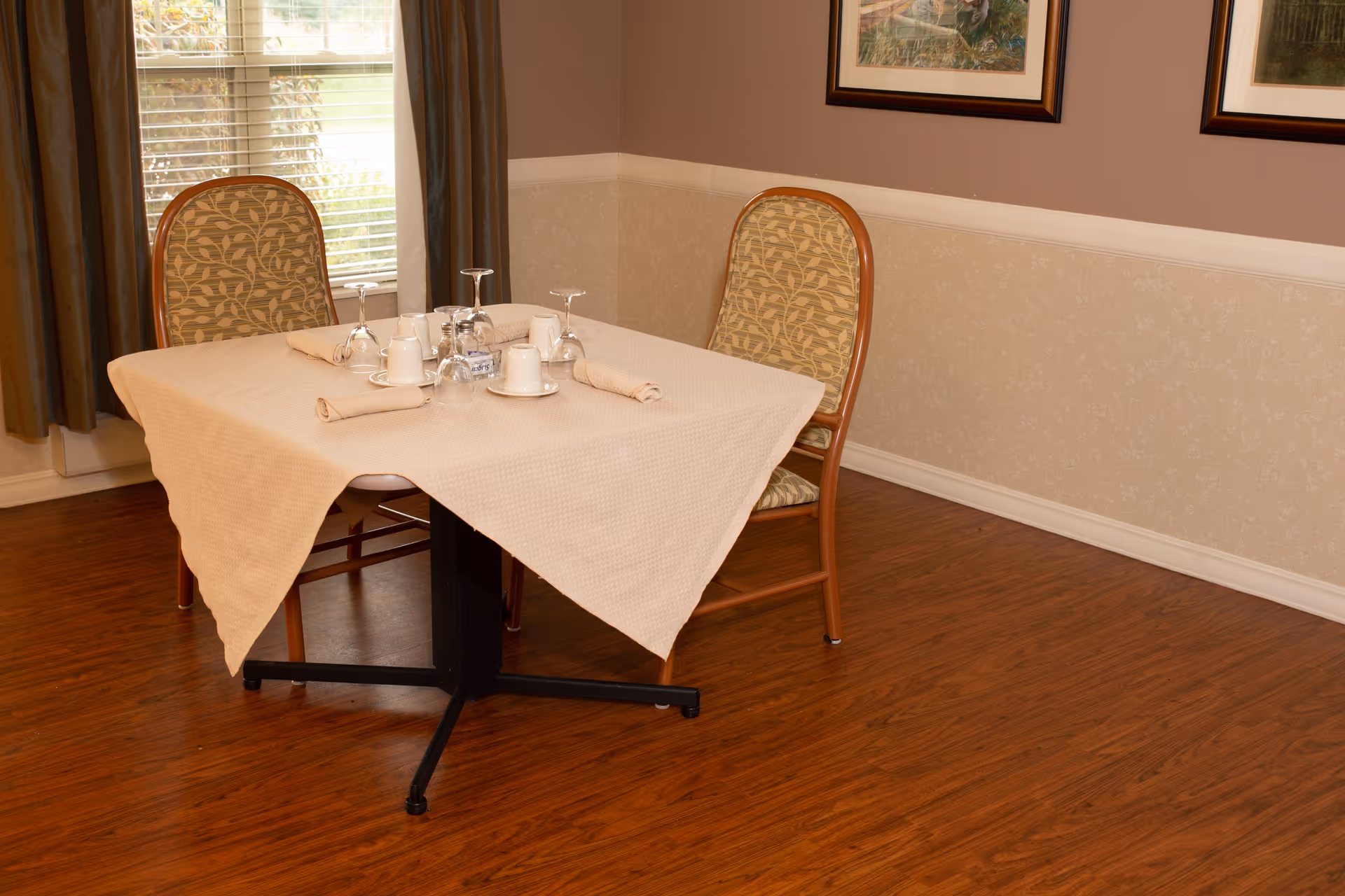 A small dining area with a square table covered by a beige tablecloth, set with upside-down wine glasses, cups, saucers, and rolled napkins. Two wooden chairs with patterned cushions are placed on either side of the table. The room has wooden flooring, beige wallpaper with a white chair rail, and a window with brown curtains letting in natural light. Two framed pictures hang on the wall.