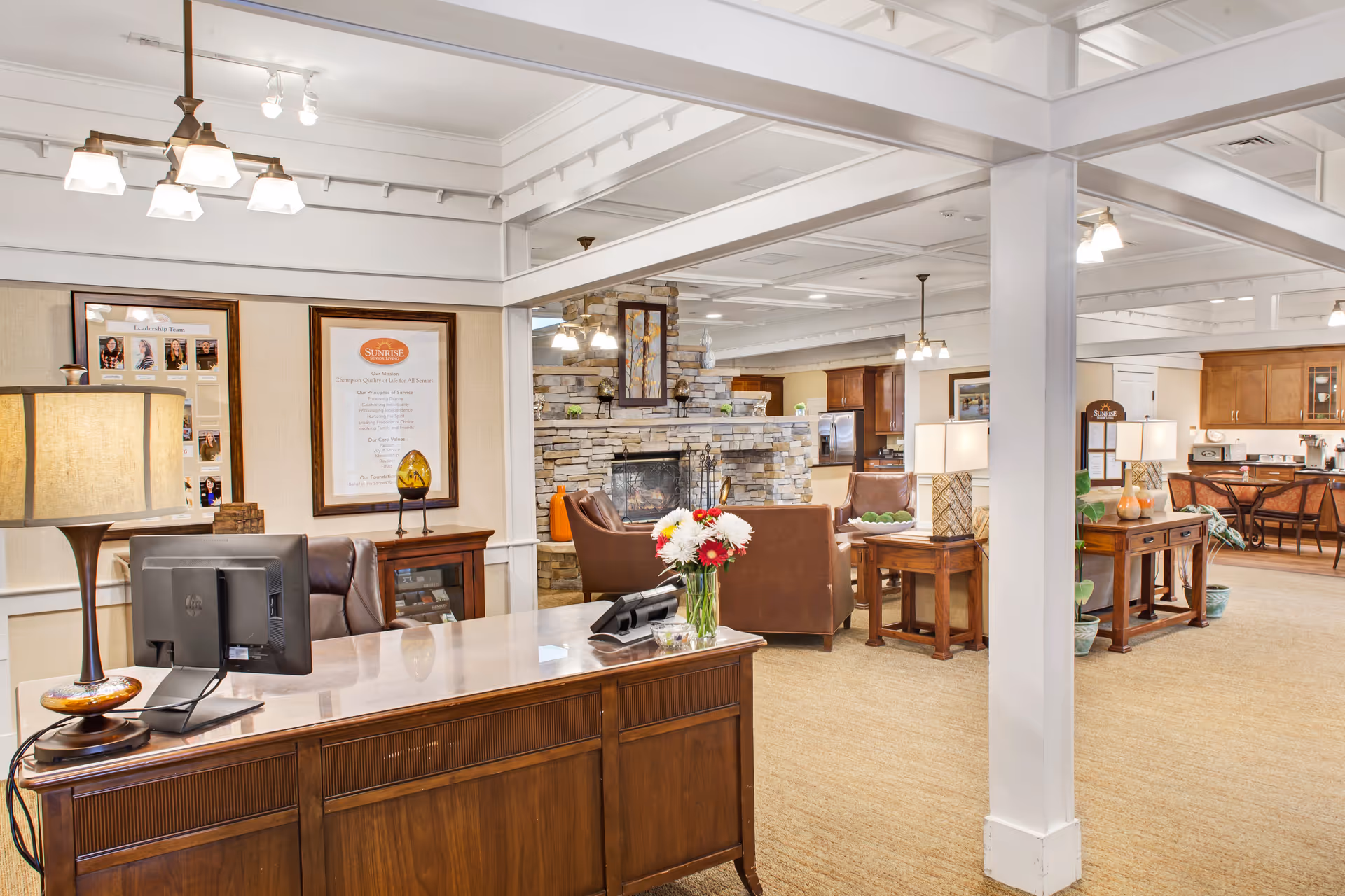 Interior view of a senior living facility reception and common area with a wooden desk and computer in the foreground, a stone fireplace surrounded by brown leather chairs in the middle, and a kitchen area with wooden cabinets in the background. The space is warmly lit with ceiling lights and table lamps, and decorated with flowers and framed pictures on the walls.