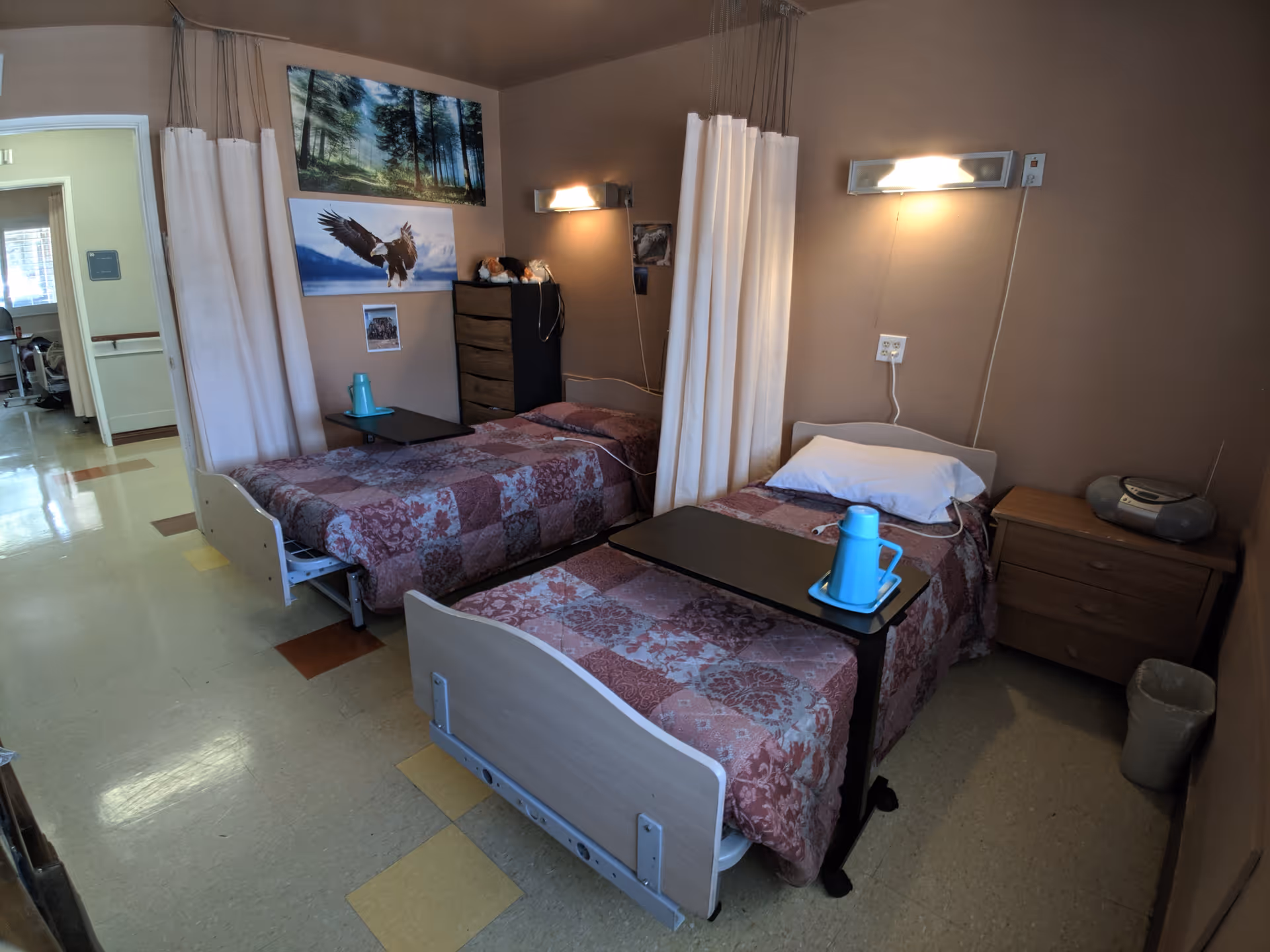 Shared senior-living bedroom with two hospital-style beds, privacy curtains, bedside tables and wall lights.