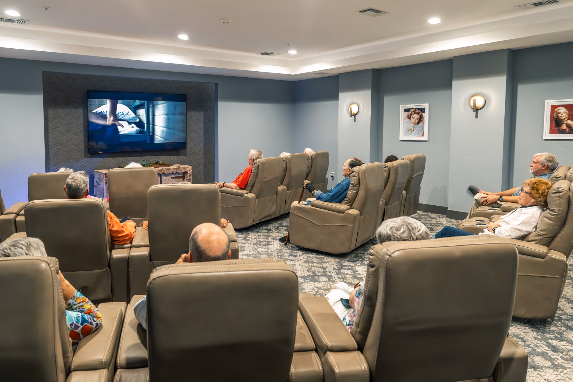 A group of elderly people seated in comfortable recliner chairs watching a movie on a large wall-mounted TV in a cozy, well-lit room with blue-gray walls and framed pictures.