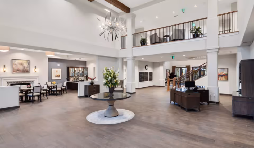 Spacious two-story senior living facility lobby with a central round table, seating and dining area, reception desk and staircase.