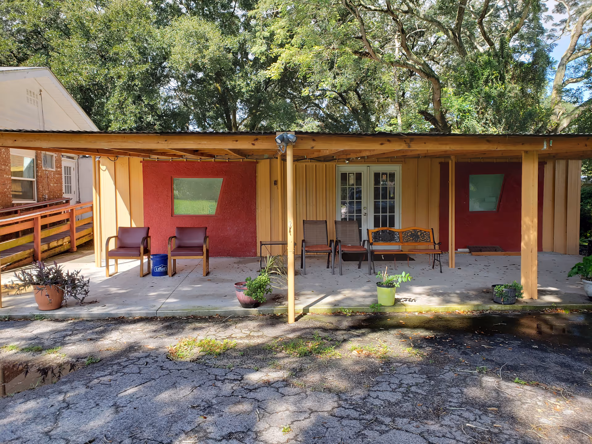 Outdoor covered patio area with several chairs and benches arranged along the wall of a building. There are potted plants placed on the concrete floor and large trees providing shade in the background.