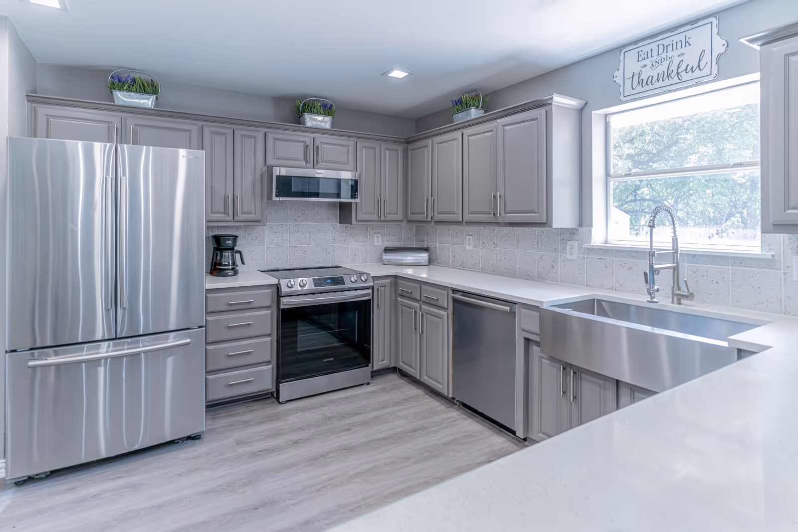 Modern kitchen with stainless steel refrigerator, oven, dishwasher, large farmhouse sink, gray cabinets and a window with a decorative 'Eat Drink and be thankful' sign.