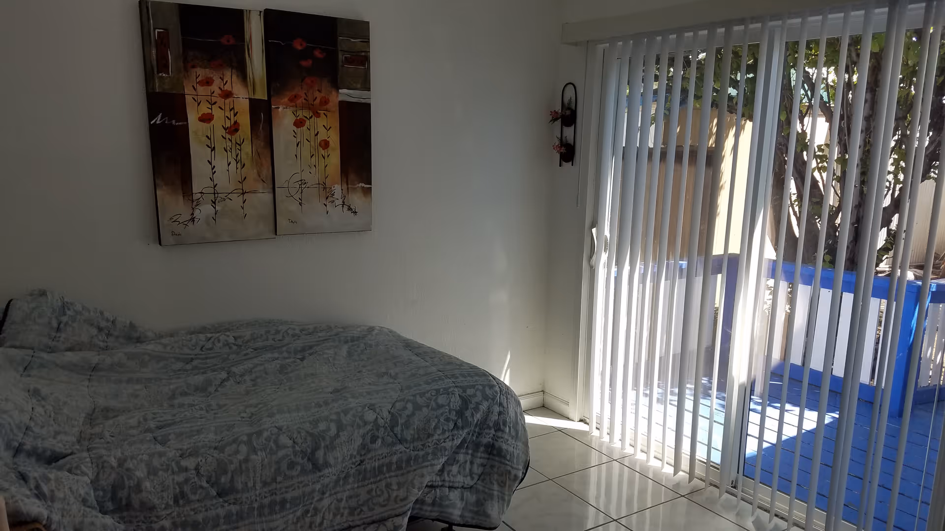 A bedroom with a bed covered in a patterned blue and gray comforter, a two-panel floral painting on the wall above the bed, and vertical blinds covering a sliding glass door that opens to a small outdoor balcony with blue railings.