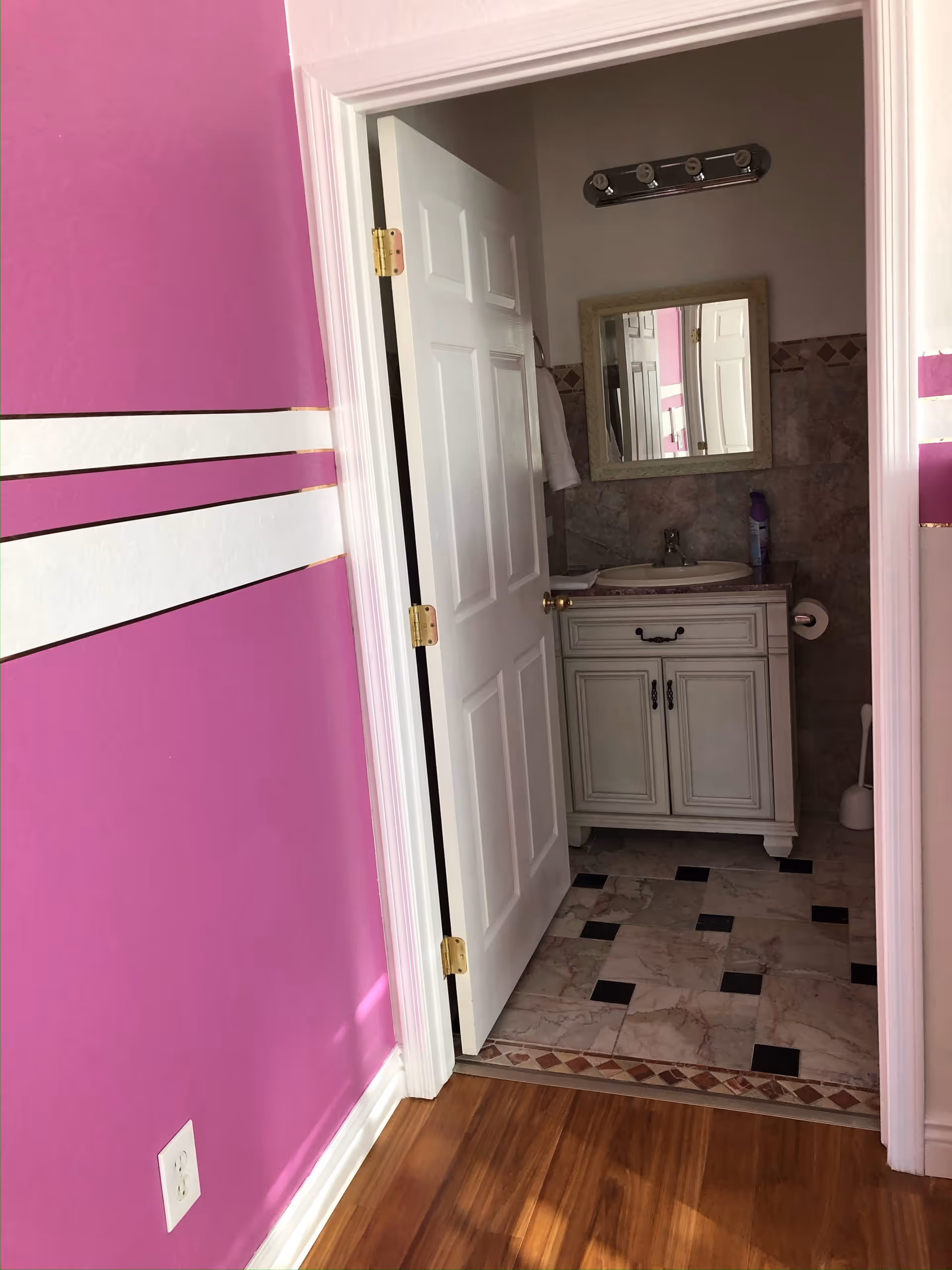 View of a bathroom through an open white door from a room with purple walls and white horizontal stripes. The bathroom has a white vanity with a sink, a mirror above it, a light fixture with four bulbs, and tiled floor and walls.