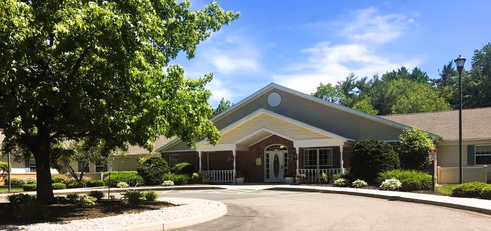 Front exterior view of a single-story assisted living facility building with a peaked roof, a covered entrance with white columns, surrounded by green trees and shrubs under a blue sky with some clouds.