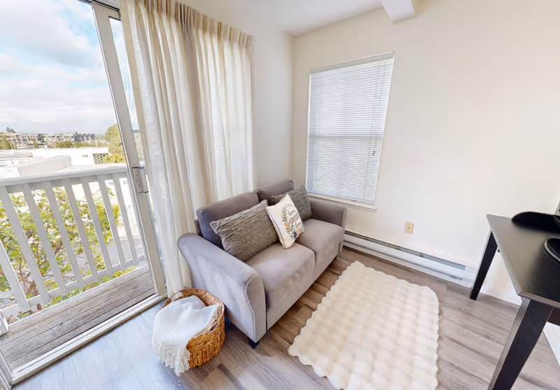 A small living room area with a gray loveseat adorned with three pillows, one with a floral design. Next to the loveseat is a wicker basket with a white blanket. A white textured rug lies on the wooden floor. There is a sliding glass door with sheer curtains leading to a balcony with a white railing, and a window with closed blinds on the adjacent wall. A black desk is partially visible on the right side.