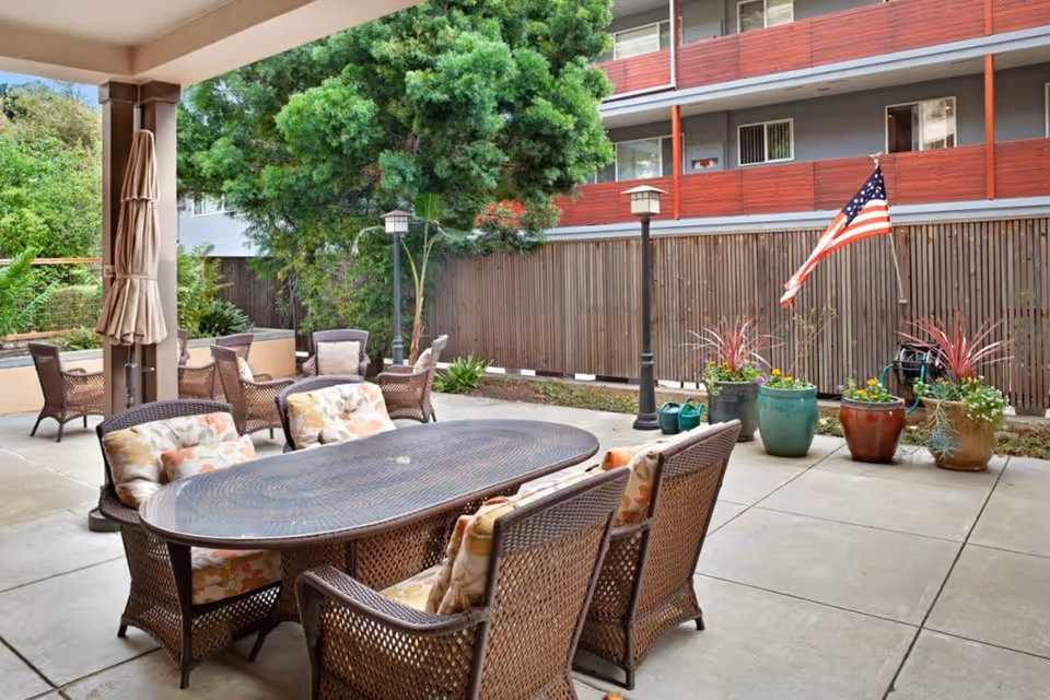 Outdoor patio area with a large oval table surrounded by wicker chairs with floral cushions. There are additional wicker chairs and tables in the background, a large tree, potted plants, an American flag, and a wooden fence with a multi-story building behind it.