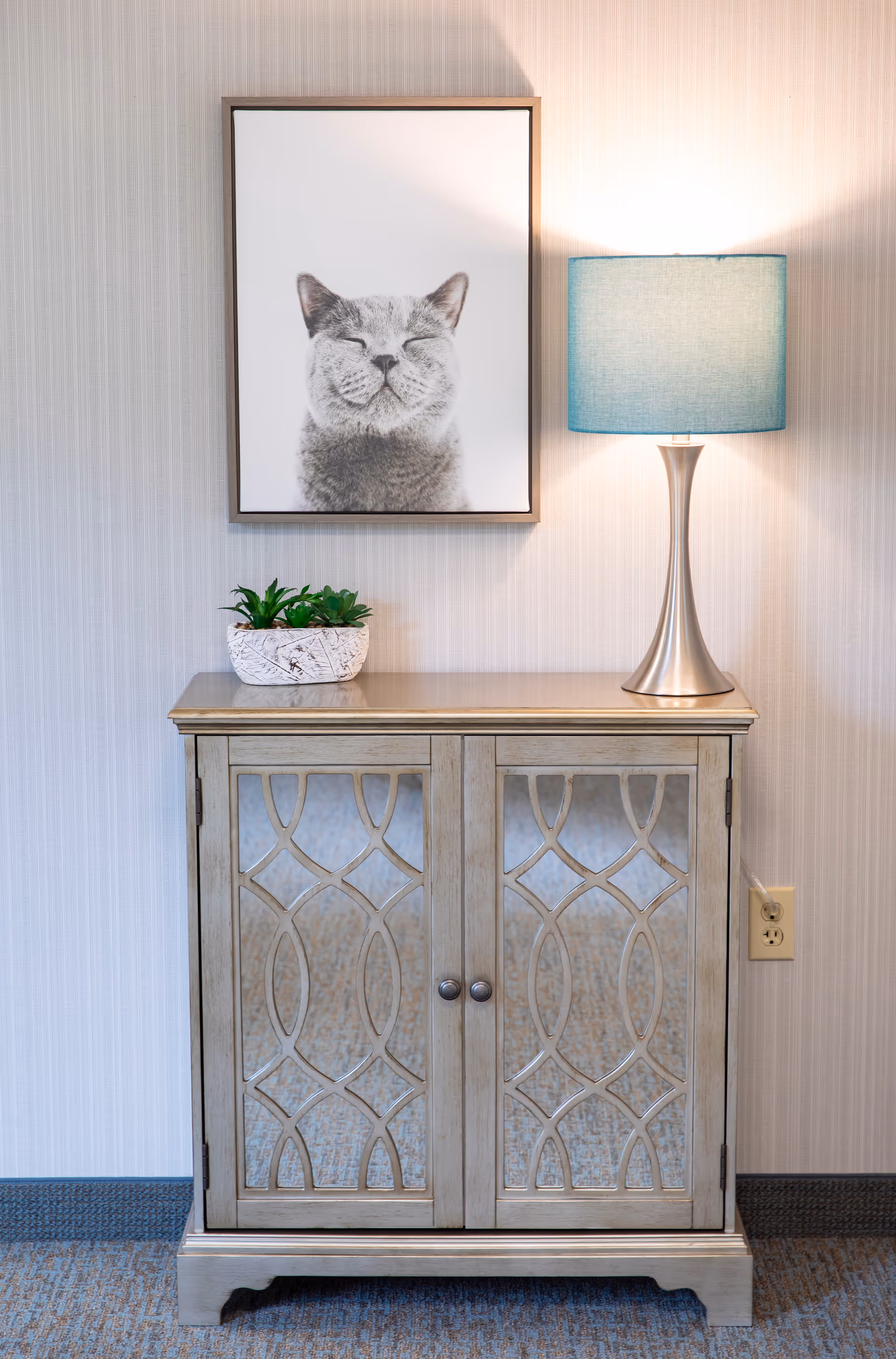A decorative wooden cabinet with glass doors featuring a geometric pattern, topped with a small potted plant and a modern table lamp with a blue lampshade. Above the cabinet hangs a framed black and white photograph of a smiling cat. The setting is indoors against a light-colored textured wall and carpeted floor.