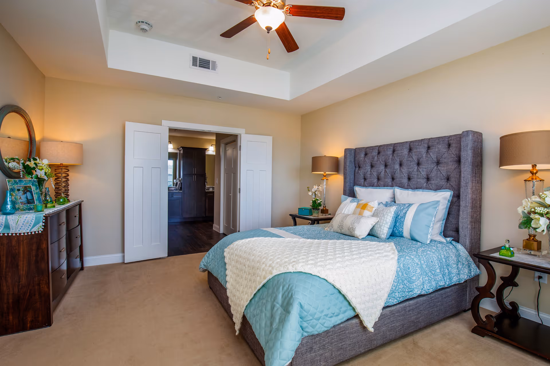 A well-lit bedroom with a large upholstered bed featuring blue and white bedding and multiple pillows. There are two bedside tables with lamps and floral decorations on each side of the bed. A wooden dresser with a round mirror and decorative items is on the left side of the room. The room has beige walls, a ceiling fan with a light, and an open doorway leading to another room with dark wood flooring.