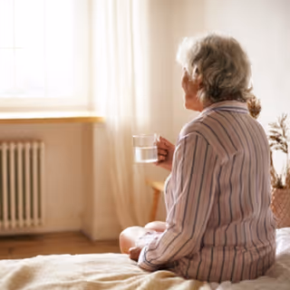 An elderly woman with gray hair wearing striped pajamas sits on a bed holding a glass of water, looking out a window with sheer curtains in a softly lit room.