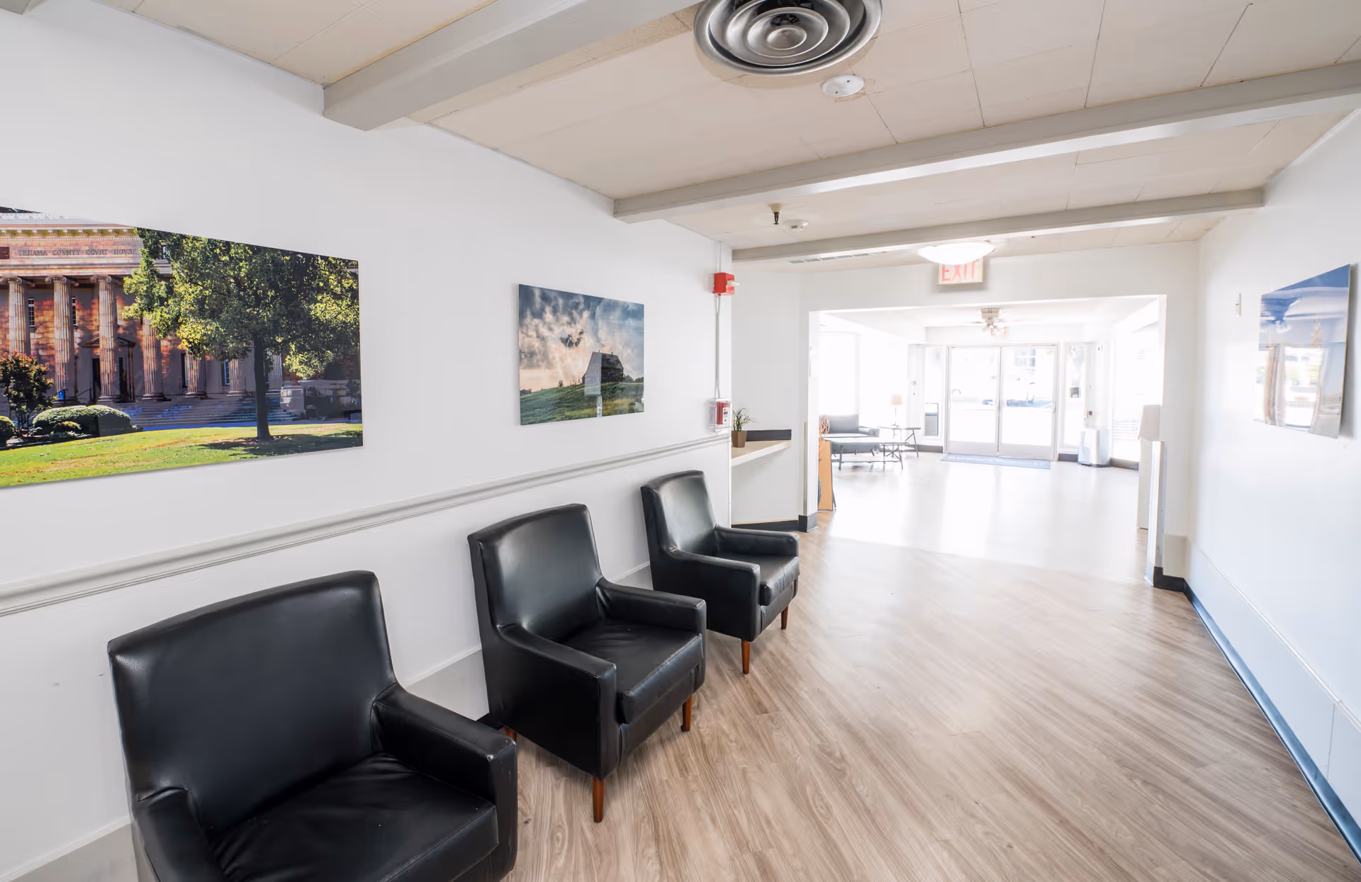 A bright hallway in a senior living facility with three black leather chairs lined up against a white wall. The walls are decorated with framed landscape photographs. The floor is light wood, and the hallway leads to a well-lit common area with large windows and glass doors.