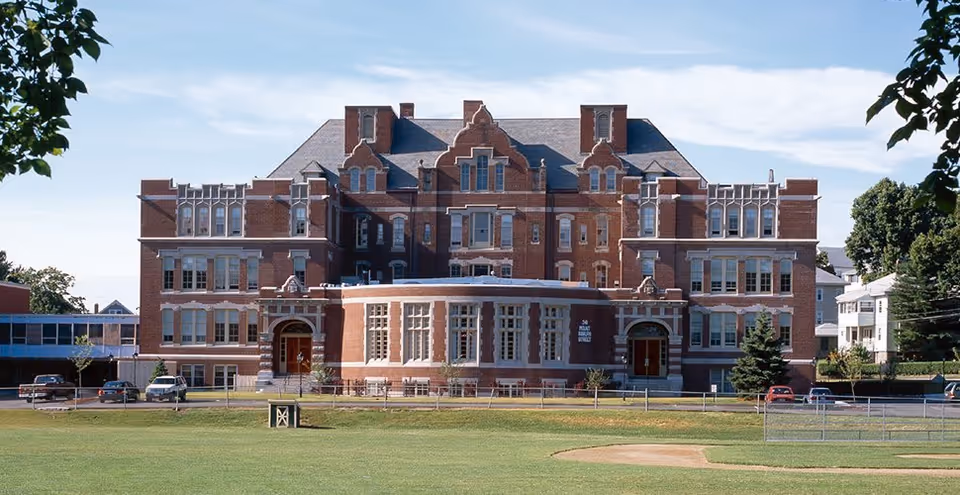 A large, historic brick building with multiple windows and decorative architectural details, situated behind a grassy field with a baseball diamond in the foreground. The sky is clear with a few clouds, and trees frame the scene on both sides.