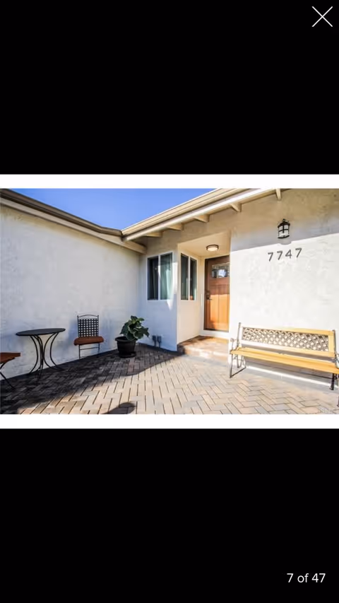 Outdoor patio area at the entrance of Ana's Residence Care Facility with a wooden bench, a small round table, two chairs, a potted plant, and the building number 7747 displayed on the wall next to a wooden door.