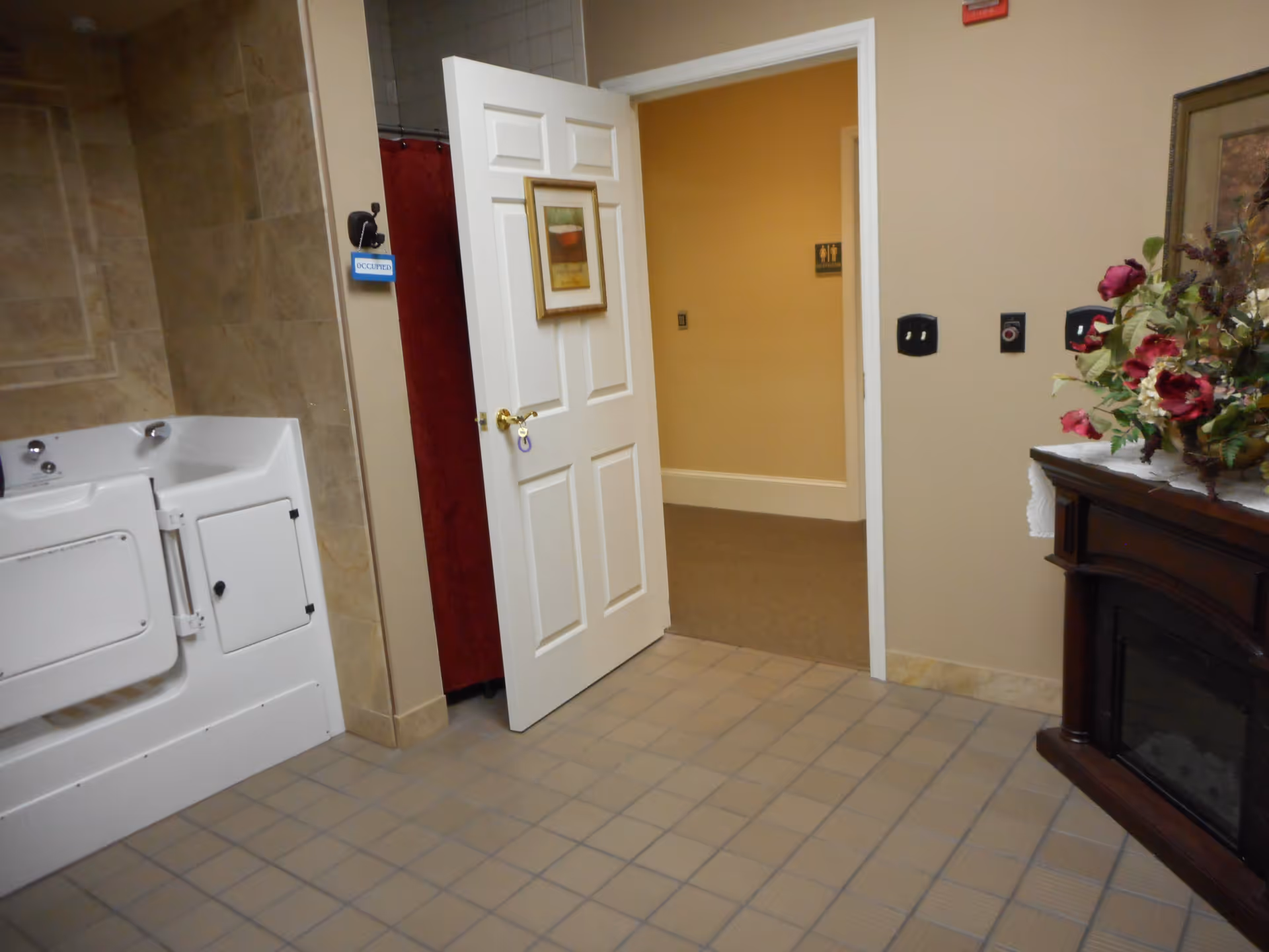Interior view of a bathroom area in Feridean Commons featuring a white walk-in bathtub with beige tiled walls and floor. A white door is open, showing a hallway with a restroom sign. To the right, there is a wooden cabinet with a floral arrangement on top.