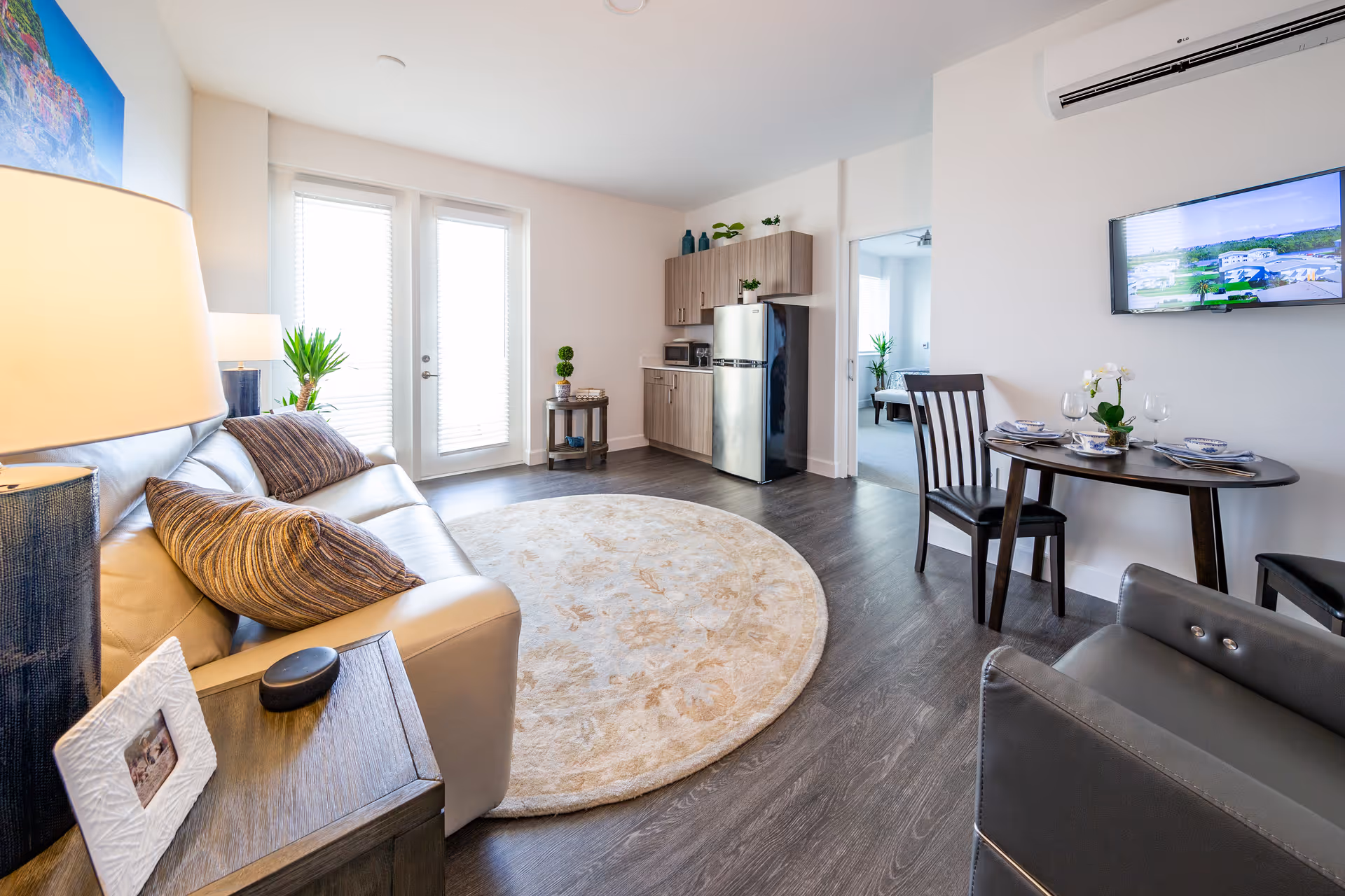 A bright and modern living area with a beige leather sofa adorned with striped pillows, a round beige patterned rug on dark wood flooring, a small dining table set for two with wine glasses, a wall-mounted TV, and a compact kitchen area with a stainless steel refrigerator and wooden cabinets. There is a door with glass panels letting in natural light and a glimpse of a bedroom through an open doorway.