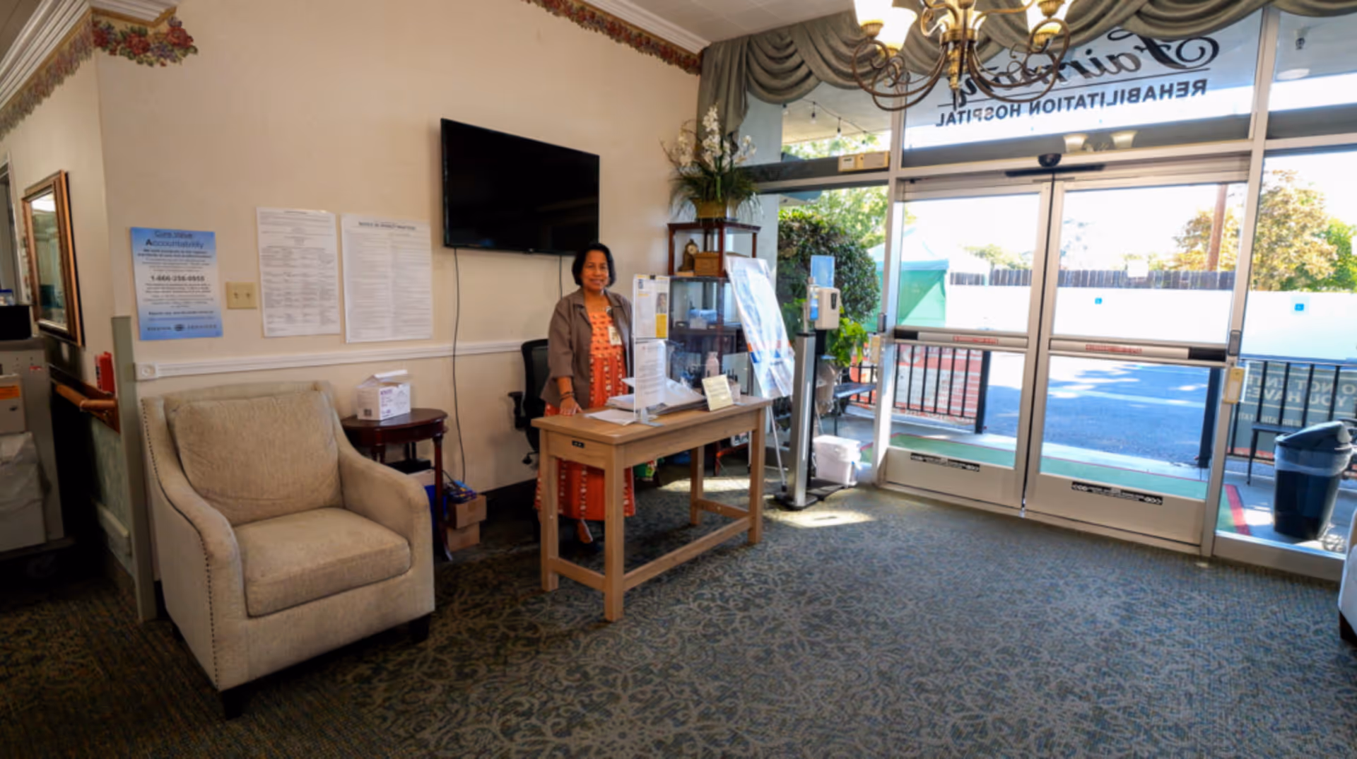 Reception area of Fairmont Rehabilitation Hospital with a woman standing behind a wooden desk. The area has a beige armchair, a wall-mounted TV, informational posters on the wall, and large glass doors leading outside. The space is carpeted and well-lit with natural light.
